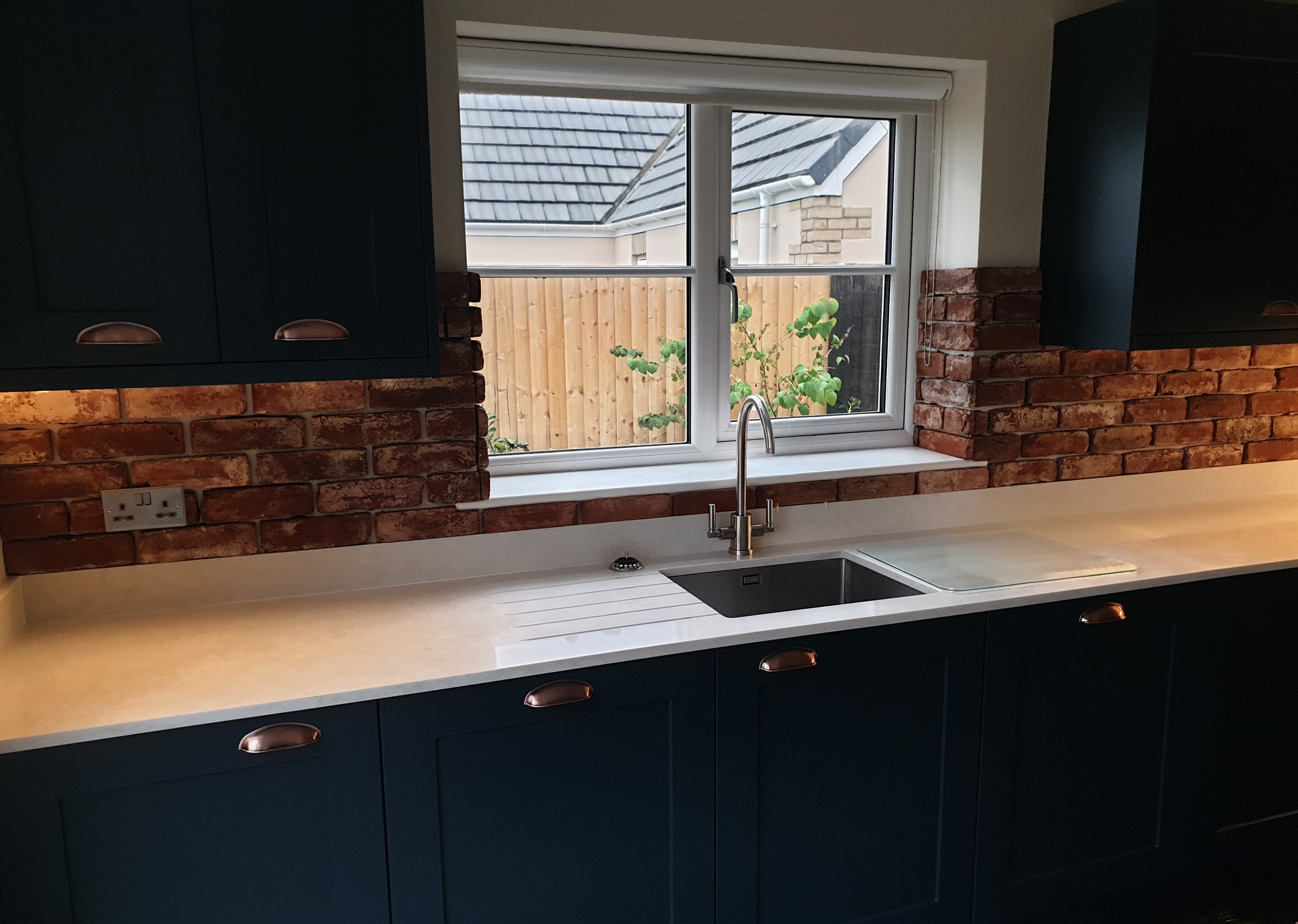 Kitchen with dark cabinetry, a white worktop and a brick-effect splashback. The sink sits beneath the window, making good use of the space.