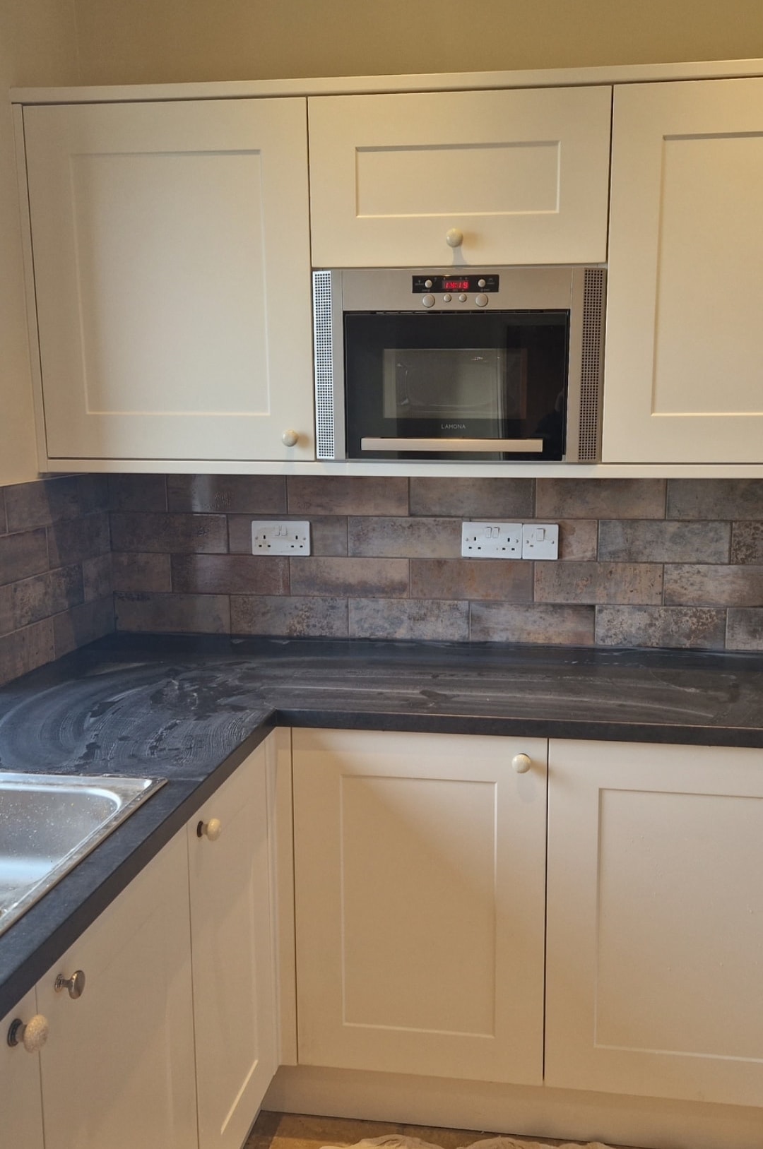 Kitchen detail showing cream cabinetry, a dark worktop and grey brick-effect splashback, with the oven built into the wall units.